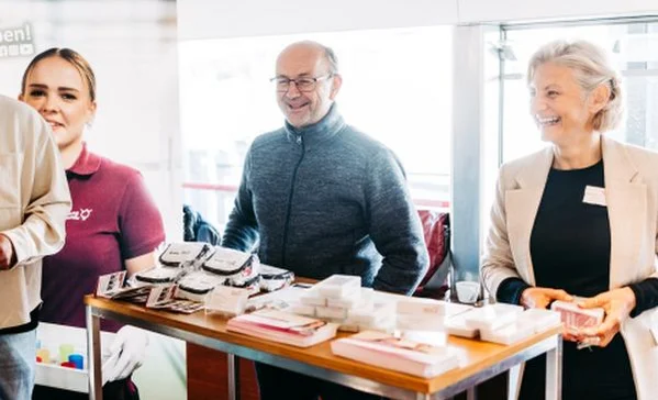 Drei lächelnde Menschen – eine junge Frau, ein älterer Mann und eine ältere Frau – stehen hinter einem Tisch mit verschiedenen kleinen verpackten Produkten und Informationsmaterialien, offenbar an einem Infostand oder Messetisch.