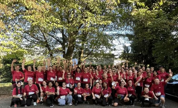 Große Gruppe von etwa 60 Personen in roten T‑Shirts steht unter Bäumen, lächelt und winkt in die Kamera – Teamfoto bei einer Outdoor‑Veranstaltung.